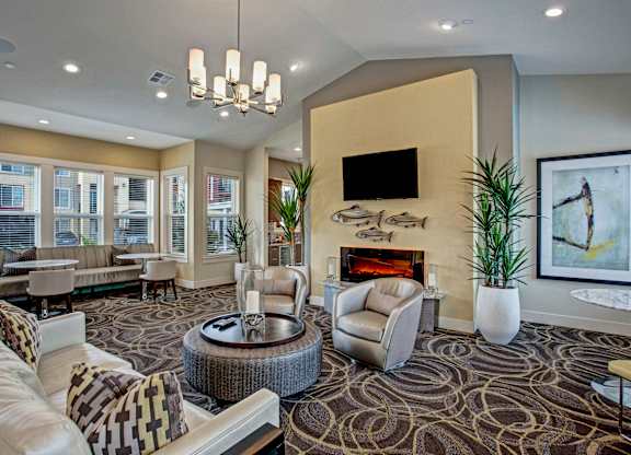 A cozy resident lounge here at Riverside 9 with beige seating, patterned carpet, modern fireplace, and large bright windows creating a warm, inviting gathering space.