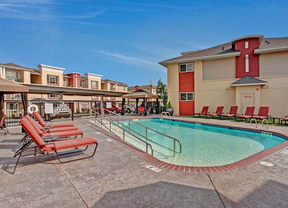 A sunny outdoor pool here at Riverside 9 with clear water, red lounge chairs, shaded seating areas, and a welcoming community setting.