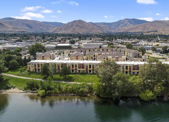 An aerial exterior view here at Riverside 9 showcasing beautiful riverfront apartment buildings, lush green landscaping, and scenic mountain views in the background.