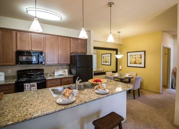 A welcoming kitchen here at Riverside 9 with a large granite breakfast bar, warm wood cabinets, black appliances, pendant lighting, and an open layout connecting to the dining area.