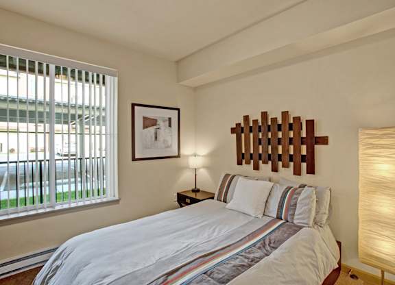A cozy bedroom here at Riverside 9 with a comfortable bed, warm wood headboard accent, bedside table with lamp, and a large window with vertical blinds bringing in natural light.