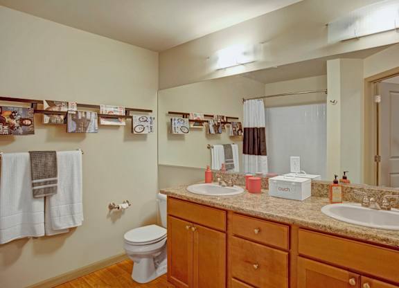 A spacious bathroom here at Riverside 9 with a dual sink vanity, large mirror, warm wood cabinetry, granite countertop, and a shower-tub combination with neutral curtain.