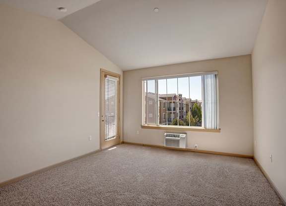A spacious, empty living room here at The Vintage with soft beige carpet, light neutral walls, and a large window bringing in natural light. A glass door with blinds leads outside, and a wall-mounted air conditioner sits below the window.