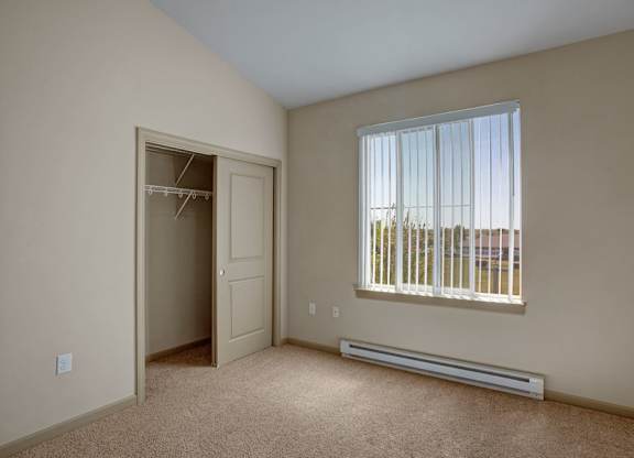 A bright bedroom here at The Vintage with soft beige carpet, light neutral walls, and a window with vertical blinds bringing in natural light. A spacious closet with sliding doors and built-in shelving provides convenient storage.