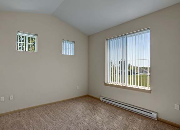 A spacious, empty bedroom here at The Vintage with soft beige carpet, light neutral walls, and multiple windows with vertical blinds bringing in natural light and open outdoor views.