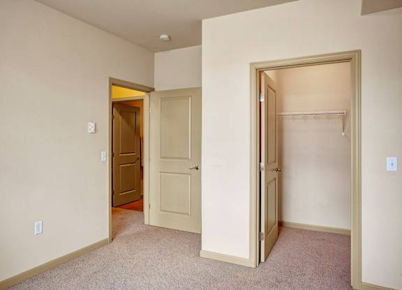 A bright bedroom here at The Vintage with soft beige carpet, light neutral walls, and an open doorway leading to the hallway. A spacious closet with built-in shelving provides convenient storage.