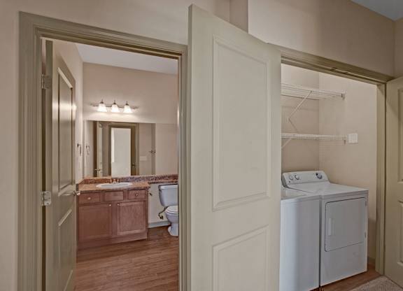 A convenient laundry area here at The Vintage with a full-size washer and dryer tucked into a closet with built-in shelving. A nearby bathroom features a wooden vanity with a speckled countertop, large mirror, and bright overhead lighting.