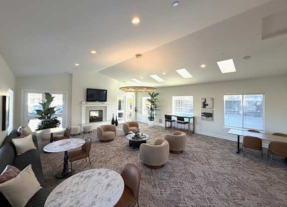 A spacious resident lounge here at The Vintage with patterned carpet, large windows bringing in natural light, and multiple seating areas with round tables and upholstered chairs. A cozy fireplace with a mounted TV and modern lighting create a welcoming gathering space.