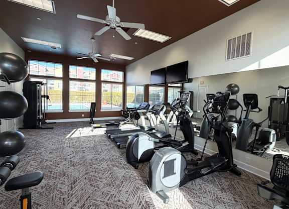 A bright fitness center here at The Vintage with large windows bringing in natural light and views of the pool area. Cardio machines, weight equipment, and exercise balls sit beneath high ceilings with fans and modern lighting.