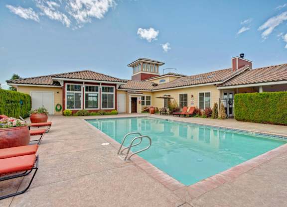 A refreshing outdoor pool here at The Vintage with clear blue water, surrounding lounge chairs with red cushions, and a spacious patio area. A clubhouse with large windows and landscaped greenery creates a relaxing resort-style setting.