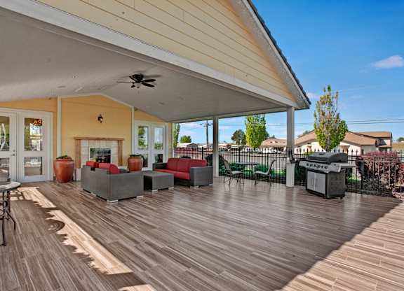 A covered outdoor patio here at The Vintage with wood-style flooring, comfortable seating with red cushions, and a cozy fireplace. A built-in grill and ceiling fan create a welcoming space for relaxing and outdoor gatherings.