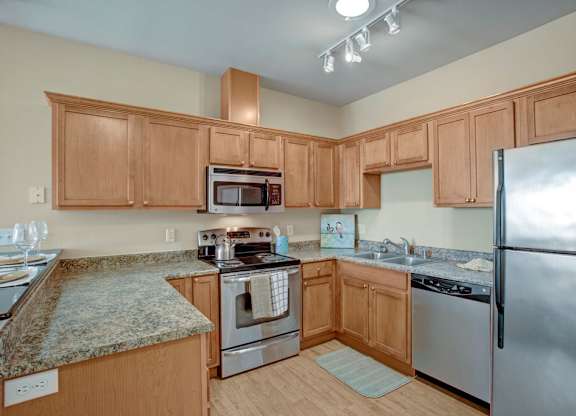 A spacious kitchen here at The Vintage with light wood cabinets, speckled granite-style countertops, and stainless steel appliances including a refrigerator, microwave, stove, and dishwasher. A double sink and bright overhead lighting create a clean, modern feel.