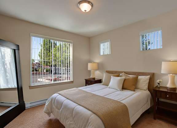 A cozy bedroom here at The Vintage with soft beige carpet, warm cream walls, and large windows with vertical blinds bringing in natural light. A bed with white bedding and neutral accent pillows sits between wooden nightstands with matching lamps.