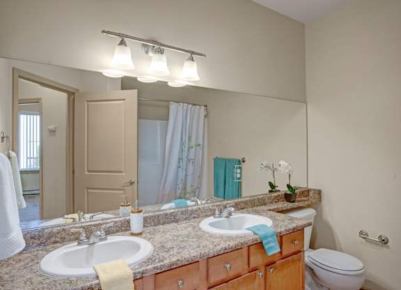 A clean bathroom here at The Vintage with a wide mirror, double sinks, and a speckled granite-style countertop. Light wood cabinets add warmth, while bright vanity lighting and a shower with a white curtain create a fresh, inviting space.