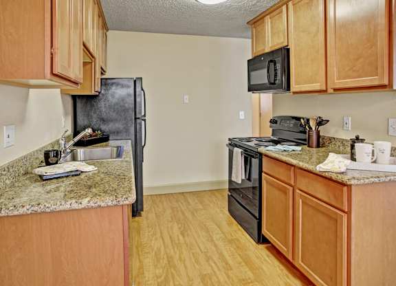 A bright kitchen here at Whispering Cedars with light brown cabinets, granite countertops, and black appliances, featuring a stainless steel sink, built-in microwave, and wood-style flooring, creating a clean, modern space with a warm, welcoming feel.