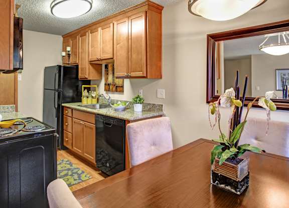 A cozy kitchen here at Whispering Cedars with light brown cabinets, granite countertops, and black appliances, featuring a stainless steel sink, dishwasher, and adjacent dining area with a wooden table and decorative mirror, creating a warm, inviting feel.