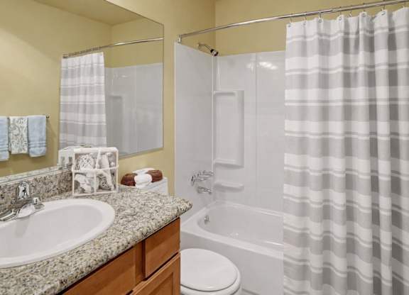A clean bathroom here at Whispering Cedars with a bathtub-shower combo, white surround, and striped shower curtain, featuring a granite countertop vanity with a large mirror, white sink, and warm wood cabinets, creating a bright, polished feel.