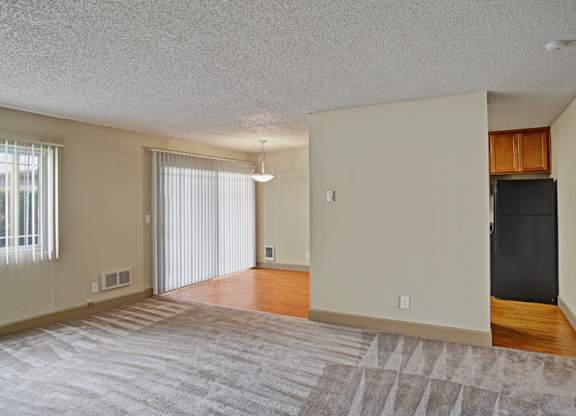 A spacious, empty living room here at Whispering Cedars with soft carpet, large windows with vertical blinds, and abundant natural light, opening to a dining area with wood-style flooring and a sliding glass door, creating a bright, open layout.