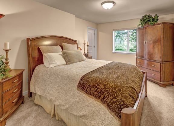 Cozy bedroom with a large wooden bed, ornate pillows, and a patterned throw blanket. Wooden dresser and plants by a window create a warm, inviting ambiance. Located at Admirals Cove Apartments.