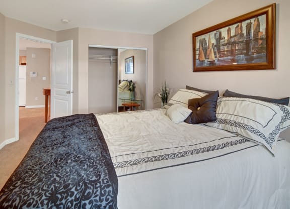 Cozy Admirals Cove Apartment bedroom with a neatly made bed featuring patterned sheets and pillows. A large painting hangs above the bed, and a closet with mirrored doors is open. Soft neutral tones create a calm ambiance.