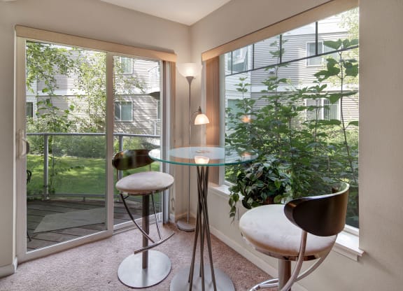 Modern dining nook with a glass table and two sleek metal stools, beside large windows overlooking lush greenery. Bright, serene atmosphere. Located at Admirals Cove Apartments.