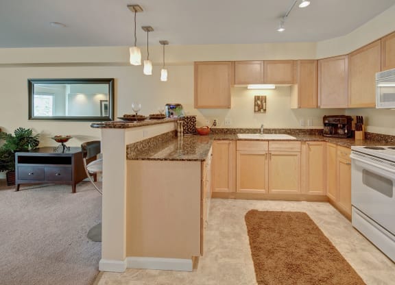 Modern kitchen with light wood cabinets, granite countertops, white appliances, and a beige rug. The kitchen opens to a living area with a mirror, small table, and decor at Admirals Cove Apartments.