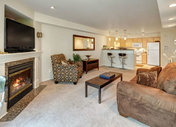 Cozy living room at Admirals Cove with a lit fireplace, wall-mounted TV, and patterned armchair. Open kitchen with bar stools and modern appliances. Warm tones.
