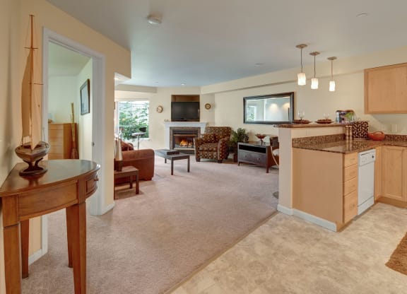 Spacious Admirals Cove living room with beige walls and carpet, featuring a cozy fireplace, brown sofa, patterned armchair, TV, and an adjacent kitchen with a granite countertop