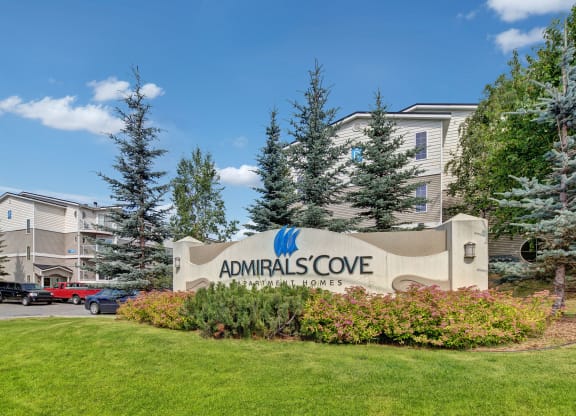 Sign for Admirals Cove Apartment Homes is surrounded by lush greenery and pine trees. Modern buildings are visible in the background under a clear blue sky.