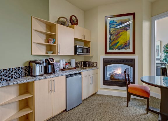 Cozy kitchen in the community lounge with light wood cabinets, granite countertop, microwave, and coffee maker. A small fridge, glass table, and lit fireplace add warmth. Located at The Alpine Apartments.