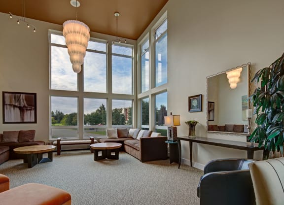Spacious community lounge with large windows, modern chandelier, and neutral tones. Features a sectional sofa, round tables, and a decorative mirror. Airy and elegant. Located at The Alpine Apartments.