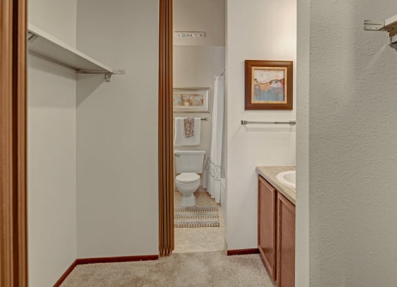 A narrow bathroom with beige walls features a toilet and sink. Artwork and a towel hang on the walls, while a striped rug adds subtle color. Located at Alpine Apartments.
