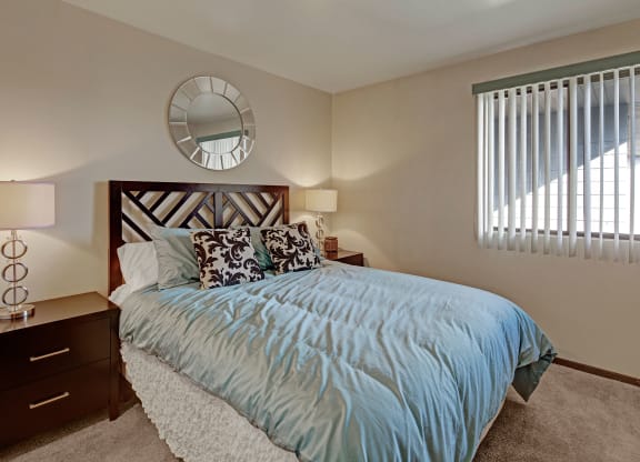 Alpine Apartments. Cozy bedroom with a large bed featuring a blue duvet and patterned pillows. Two bedside tables have lamps. A circular mirror hangs on the wall.