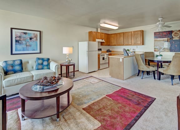 A cozy living and kitchen area with beige walls and carpet. A white sofa hosts patterned cushions beside a round table with a lamp. The kitchen features wood cabinets, and a dining area with a round table and tan chairs. A colorful abstract rug and artwork add warmth. Located at Alpine Apartments.