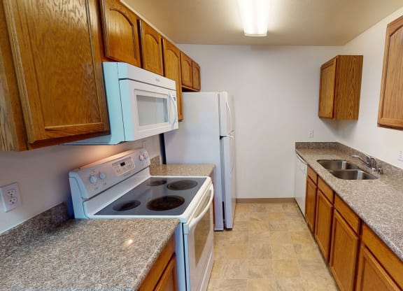 Narrow kitchen with brown cabinets, granite countertops, white stove, microwave, and fridge, and a double sink with a neutral tone. Amber Ridge Apartments.