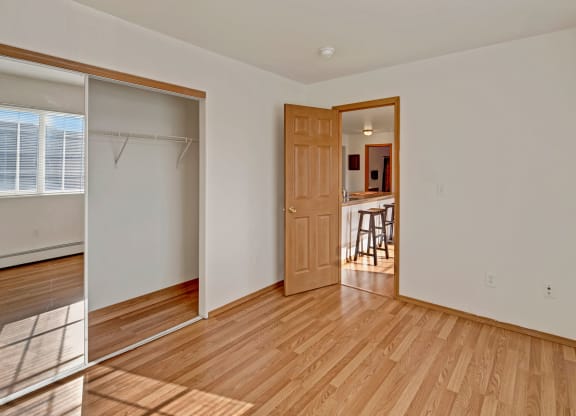 Empty bedroom with light wood flooring, white walls, and a mirrored closet with a sliding door. An open wooden door reveals a kitchen with stools. Sunlight streams through a window, creating a warm, inviting atmosphere. Amber Ridge Apartments.