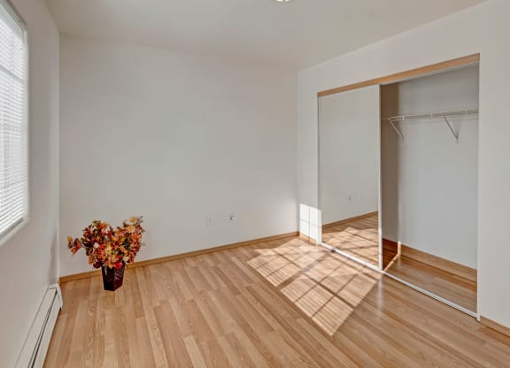 Minimalist empty bedroom with hardwood floor and white walls. A large window casts sunlight, highlighting a flower arrangement in a vase. Peaceful ambiance. Amber Ridge Apartments.