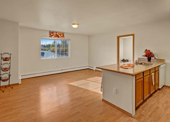 Spacious room with wooden floor, a large window, and a kitchen counter. Warm sunlight creates a cozy feel. Decorations include a floral arrangement. Amber Ridge Apartments.
