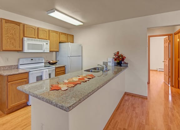 Modern kitchen with light wood cabinets, stone countertop, white appliances, and fall-themed decorations. Bright lighting and wood flooring add warmth. Amber Ridge Apartments.