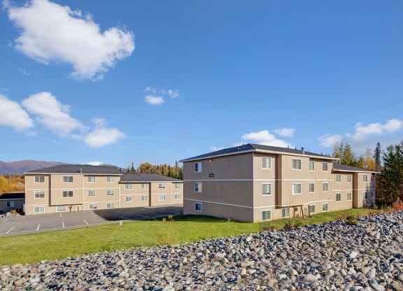 Three beige apartment buildings under a clear blue sky, surrounded by trees with autumn foliage and mountains in the distance, convey tranquility. Amber Ridge Apartments.