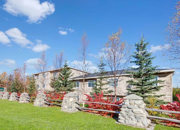Stone pillars and wooden fence enclose a building surrounded by vibrant fall trees under a bright blue sky, conveying a serene, autumnal atmosphere. Amber Ridge Apartments.