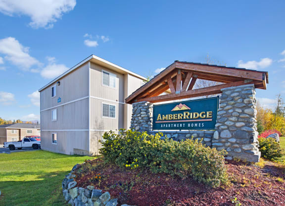 Sign for "Amber Ridge Apartment Homes" on a stone structure under a clear blue sky. Nearby are beige apartment buildings and a parked truck.