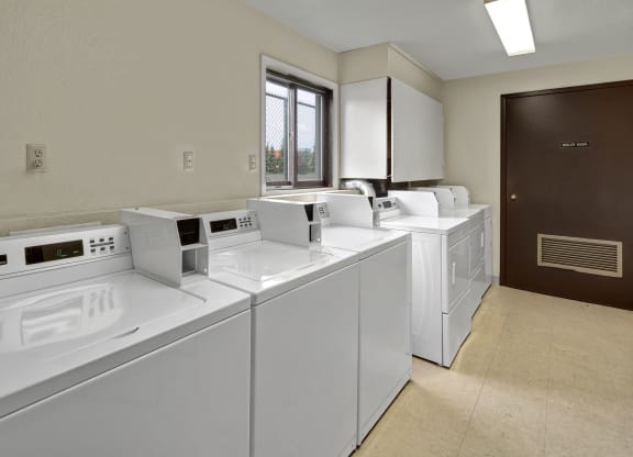 A clean laundromat with four white top-load washing machines lined up. A small window and dark door labeled "Boiler Room" are visible. Bright, orderly atmosphere. Arbor Pointe Apartments.