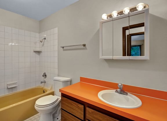 Bathroom with yellow bathtub, white tiles, and orange countertop. Includes a white toilet, mirror with lights, and wooden cabinets. Bright and vintage. Arbor Pointe Apartments.
