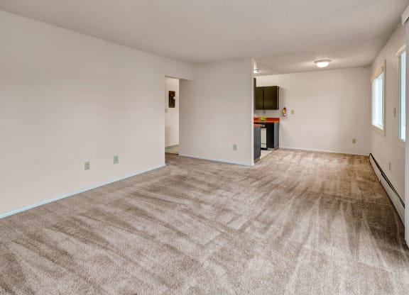 A spacious, neutral-toned living room with beige carpeting, white walls, and an open layout leading to a kitchen. Natural light filters through large windows. Arctic Sun Apartments.