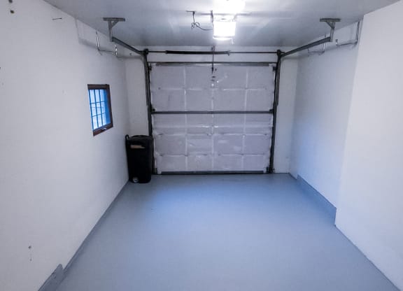 Empty garage here at Birchwood Apartments with white walls and light gray floor, bright overhead light, closed garage door at the back, and a small window on the left. Minimalistic and clean.
