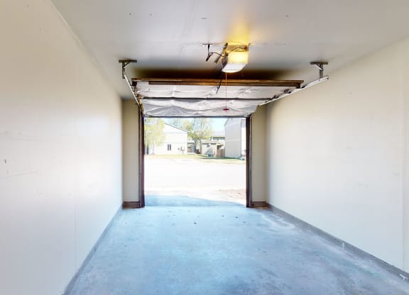 Empty garage interior here at Birchwood Homes Apartments with an open door showing a clear view of a sunny day outside. The space is clean, lit by a single ceiling light.