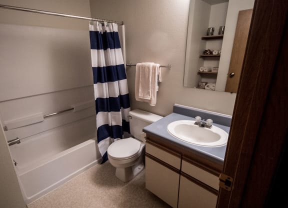 Compact bathroom here at Birchwood Apartments with a blue and white striped shower curtain, a sink with a blue countertop, toilet, towel rack, and shelf with folded towels. Clean and neat.