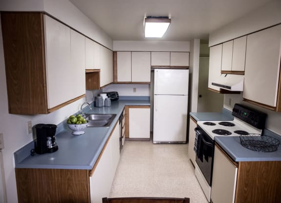 A narrow kitchen here at Birchwood Apartments with white cabinets, blue countertops, a refrigerator, stove, and sink. A bowl of green apples and a coffee maker are visible.