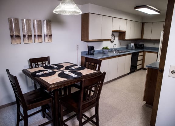 A cozy kitchen here at Birchwood Apartments with dark wooden dining table set for four, matching chairs, modern art on the wall, and a tidy countertop with fruit bowl and appliances.